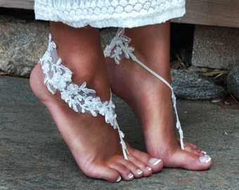 May include: White lace barefoot sandals with a delicate floral design. The sandals are tied around the ankle and have a ribbon that wraps around the toes.
