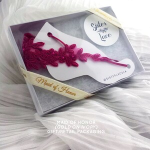 May include: A white gift box with a pink lace garter inside. The box is tied with a ribbon that says "Maid of Honor". The lid of the box has a white circle with the text "Sister Lace" in gold lettering. The box is on a white fabric background.