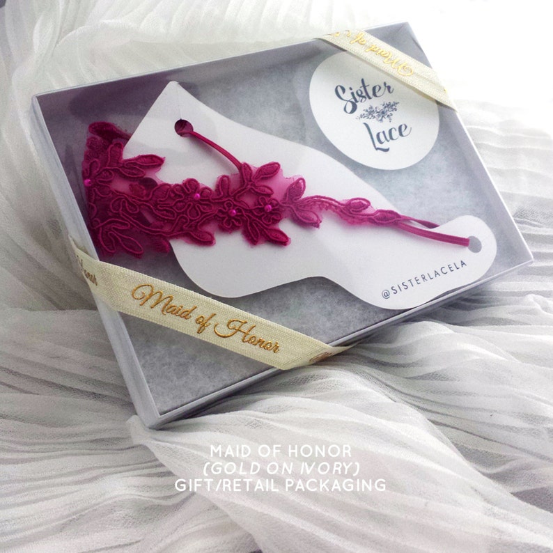 May include: A white gift box with a pink lace garter inside. The box is tied with a ribbon that says "Maid of Honor". The lid of the box has a white circle with the text "Sister Lace" in gold lettering. The box is on a white fabric background.