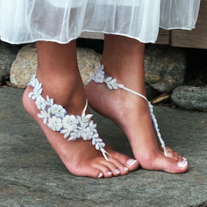 May include: White lace barefoot sandals with delicate floral details. The sandals are tied around the ankle and have a simple design.