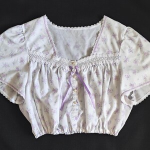 May include: A white crop top with a lavender floral print. It has a square neckline, lace trim, and short sleeves. A lavender ribbon and pearl buttons adorn the front of the garment.