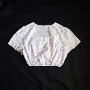 Op de afbeelding: Een witte crop top versierd met een subtiele paarse bloemenprint. Het heeft korte mouwen en een geschulpte rand op de halslijn en mouwen. De taille is gerimpeld, wat een getailleerde vorm geeft. De top is gemaakt van een lichte stof.