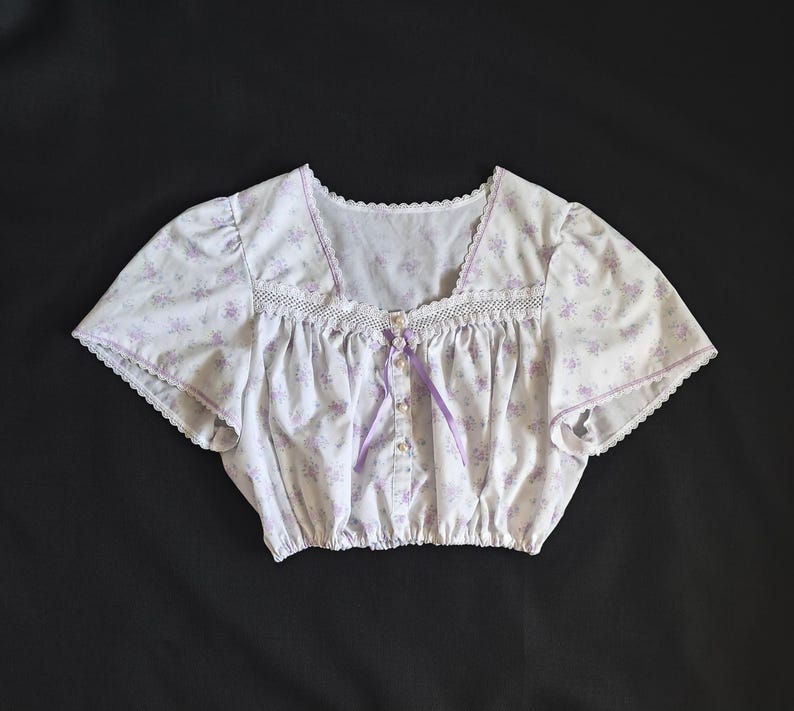 May include: A white crop top with a lavender floral print. It has a square neckline, lace trim, and short sleeves. A lavender ribbon and pearl buttons adorn the front of the garment.