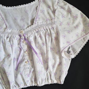 Op de afbeelding: Witte top met bloemenprint, vierkante halslijn, kanten rand en parelmoer knopen. De top heeft een delicaat patroon van paarse en blauwe bloemen, een paars lint en korte mouwen.