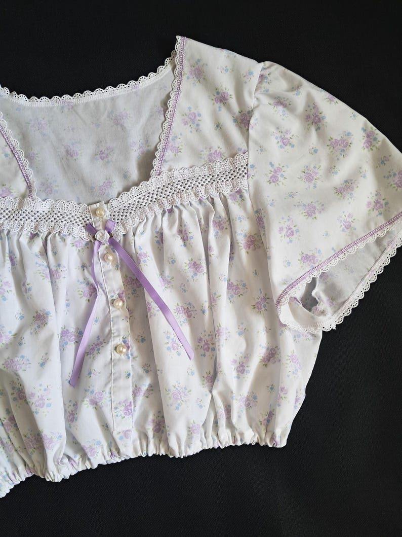 Op de afbeelding: Witte top met bloemenprint, vierkante halslijn, kanten rand en parelmoer knopen. De top heeft een delicaat patroon van paarse en blauwe bloemen, een paars lint en korte mouwen.