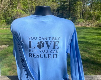 May include: Light blue long-sleeve shirt with the text "YOU CAN'T BUY LOVE BUT YOU CAN RESCUE IT" in black. The sleeve has the text "RESCUE LOVE" and a paw print. The shirt is displayed on a mannequin.