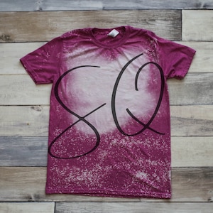 May include: A magenta t-shirt with a bleached design and the letters "SQ" in black script. The shirt has a crew neck and short sleeves. The bleached area is white and covers most of the front of the shirt.