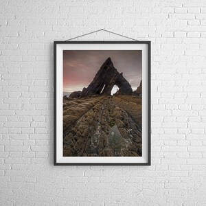 May include: A framed photograph of a rock formation with a natural archway. The rock is a dark brown colour and the sky is a light pink and purple. The rock formation is on a beach with brown sand and rocks.