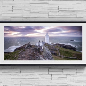 May include: A framed panoramic print featuring a white lighthouse on a rugged coastline. The lighthouse, with its tall, cylindrical structure, is set against a dramatic sky and ocean. A smaller building is adjacent to the lighthouse. The artwork is mounted on a white brick wall.