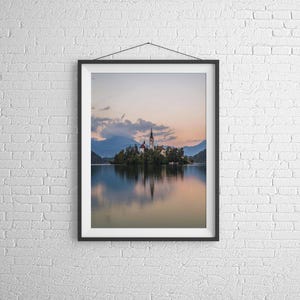 Puede incluir: Impresi&oacute;n enmarcada de un paisaje de una iglesia en una isla en un lago, con monta&ntilde;as al fondo. La imagen presenta una escena serena con reflejos en el agua y una paleta de colores suaves y pastel. La impresi&oacute;n se exhibe en una pared de ladrillo blanco.