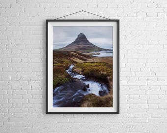 May include: A framed photograph of a mountain with a stream flowing in the foreground. The mountain is a dark gray color and the stream is a light blue color. The background is a cloudy sky.
