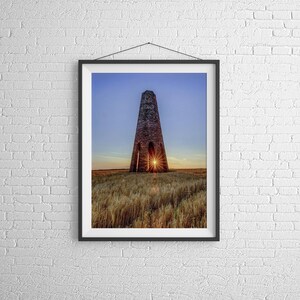May include: A tall, stone tower with a large opening at the top, silhouetted against a sunset sky. The sun is setting behind the tower, creating a bright orange glow in the sky and a starburst effect through the opening.