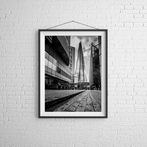 May include: A black and white photograph of the Shard, a tall skyscraper in London, England. The building is seen from a street level perspective, with other buildings in the background.