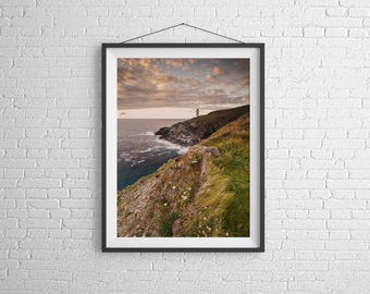 Könnte beinhalten: Ein Foto eines Leuchtturms auf einer felsigen Klippe mit Blick auf das Meer. Der Himmel ist in leuchtenden Orange- und Rosatönen gehalten, mit Wolken, die Schatten auf das Wasser werfen. Der Leuchtturm ist weiß und ragt hoch über die dramatische Landschaft.