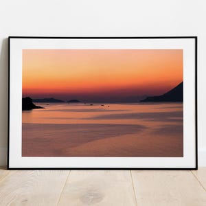 May include: A scenic photograph of a calm body of water with a mountain range in the distance. The sky is a vibrant orange and pink, creating a beautiful sunset.