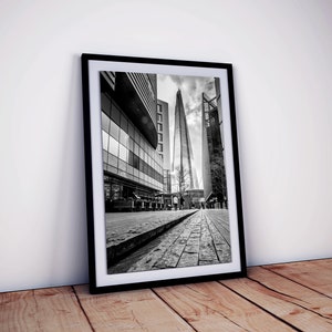 May include: Black and white photograph of a tall skyscraper in London, England. The building is surrounded by other buildings and a cobblestone street.