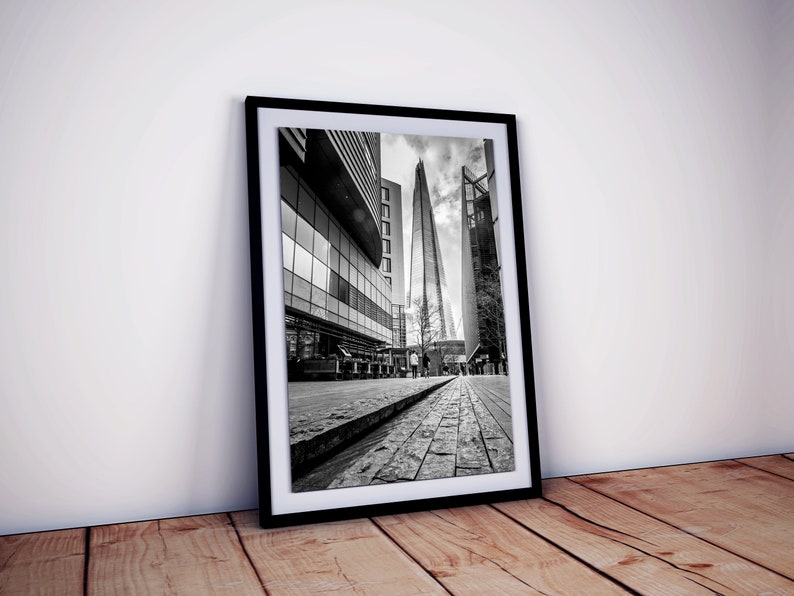 May include: Black and white photograph of a tall skyscraper in London, England. The building is surrounded by other buildings and a cobblestone street.