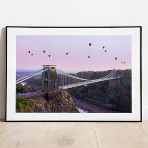 Puede incluir: Impresión enmarcada del puente colgante de Clifton en Bristol, Inglaterra, con globos aerostáticos en el cielo. El puente es de piedra gris y el cielo tiene un degradado de rosa y morado. Los globos son de varios colores.