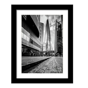 May include: A black and white photograph of a tall, modern skyscraper in London, England. The building is surrounded by other buildings and a paved street.