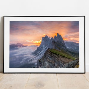 Puede incluir: Una vista panorámica de una cordillera al amanecer. El cielo es una mezcla vibrante de rosa, naranja y morado, con nubes que proyectan largas sombras sobre los picos. Las montañas son escarpadas y rocosas, con un toque de vegetación verde.