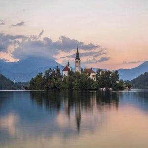 Puede incluir: Un paisaje pintoresco con una iglesia en una isla en un lago. La iglesia tiene un alto campanario y est&aacute; rodeada de &aacute;rboles. El agua refleja el cielo y la isla, creando una imagen sim&eacute;trica. Las monta&ntilde;as son visibles en el fondo bajo un cielo nublado.