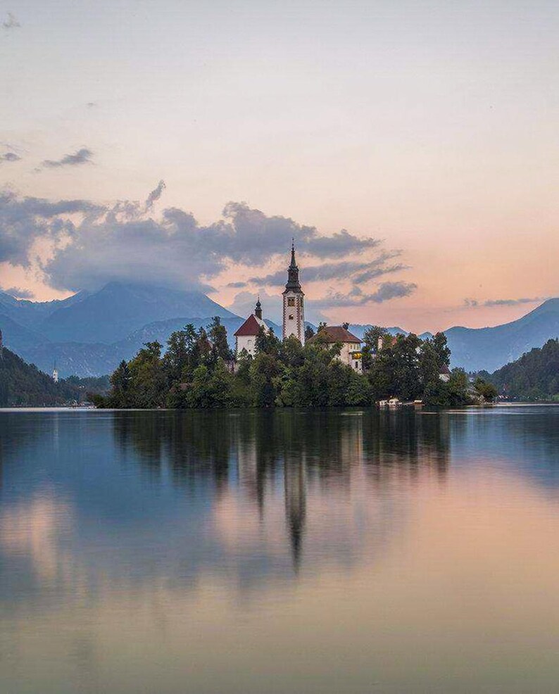 Puede incluir: Un paisaje pintoresco con una iglesia en una isla en un lago. La iglesia tiene un alto campanario y est&aacute; rodeada de &aacute;rboles. El agua refleja el cielo y la isla, creando una imagen sim&eacute;trica. Las monta&ntilde;as son visibles en el fondo bajo un cielo nublado.