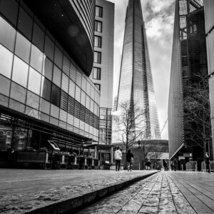 May include: A black and white photo of a modern city street with tall buildings. The buildings are made of glass and steel. There are people walking on the street. The photo is taken from a low angle, looking up at the buildings.