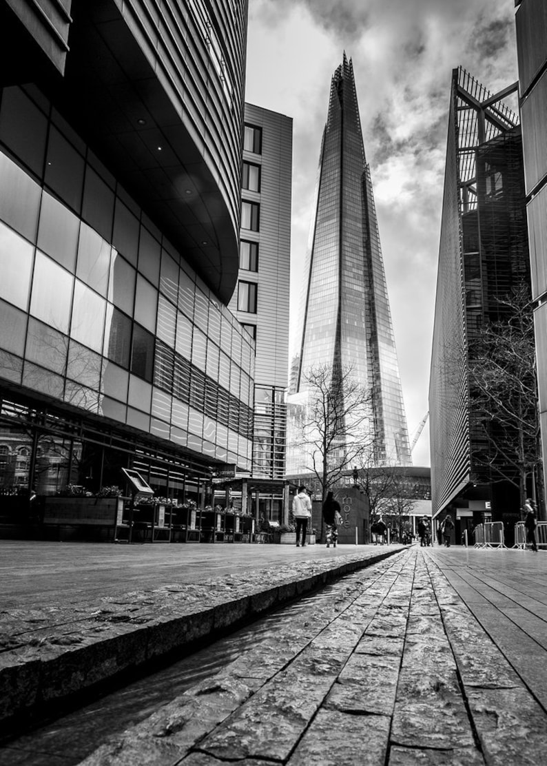 May include: A black and white photo of a modern city street with tall buildings. The buildings are made of glass and steel. There are people walking on the street. The photo is taken from a low angle, looking up at the buildings.