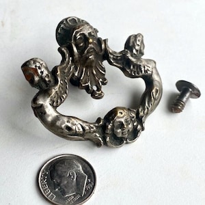 May include: Antique silver-toned metal cabinet door pull with a design of two cherubs and a bearded man's face. The pull is in the shape of a ring and has a screw for attachment.