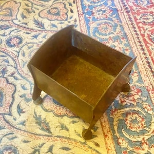 May include: A rectangular brass container with a weathered patina, featuring four curved legs and an open top. The metal surface shows signs of oxidation and age, with a deep golden-brown hue throughout the structure of this small, hollow vessel.