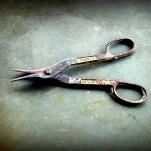 May include: A rusty metal pair of shears with a curved blade and a handle with the word "FORGED" stamped on it.