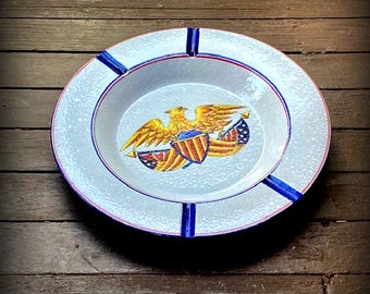 May include: A white ceramic ashtray with a blue and red rim. The center features a painted image of a golden eagle with outstretched wings holding a shield and American flags.
