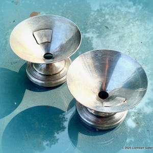 May include: Two silver metal bowls with a funnel-shaped opening in the center. The bowls are on a pedestal base with a fluted design. The bowls are on a blue surface.