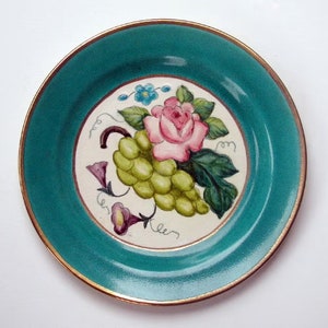 May include: A teal ceramic plate with a gold rim features a hand-painted design of a pink rose, green grapes, and blue flowers.