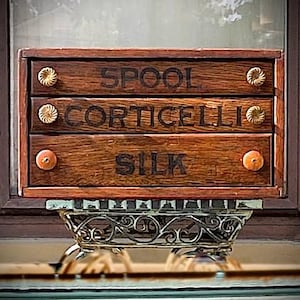 May include: A rectangular wooden spool cabinet with three drawers featuring the black text SPOOL, CORTICELLI, and SILK. Each drawer has two small round knobs. The cabinet is supported by an ornate, decorative metal base with cut-out patterns.