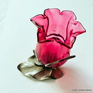 May include: A vibrant, ruby-red glass flower-shaped vase with a ruffled top. The vase is mounted on a silver-colored metal leaf and stem base. A decorative piece for home decor.