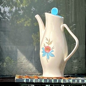 May include: A white ceramic teapot with a blue and pink floral design. The teapot has a blue lid and a handle. It is sitting on a mosaic tile surface.