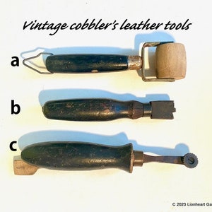 May include: Vintage cobbler's leather tools, labeled a, b, and c. Tool a is a wooden roller with a black handle. Tool b is a black handled tool with a metal edge. Tool c is a black handled tool with a metal wheel.