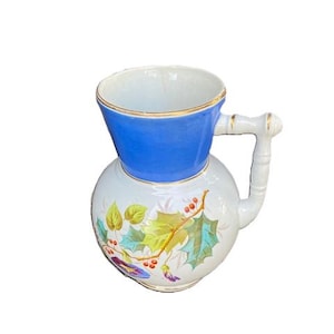 May include: A vintage white ceramic pitcher with a blue top and gold trim. The pitcher features a round body with a handle and is decorated with colorful floral and leaf designs. A classic piece for serving.