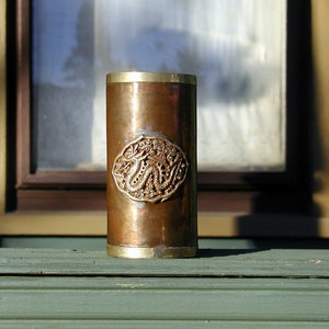 May include: A copper and brass cigarette lighter with an ornate design. The design features a stylized floral pattern with a central motif of two intertwined dragons.
