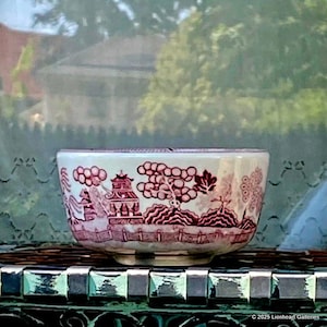 May include: A white and red ceramic bowl with a Willow pattern design. The bowl features a detailed landscape scene with pagodas, trees, and figures. The bowl is set on a decorative surface with a blurred background.