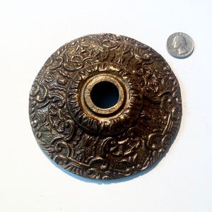 May include: A circular, dark bronze-toned metal canopy plate featuring an intricate, raised scrollwork pattern and a central opening. A United States quarter coin is positioned to the upper right for scale, showing the relative size of the piece.