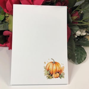 May include: A white notepad with a watercolor illustration of two pumpkins and fall leaves. The pumpkins are orange and the leaves are brown, yellow, and green.