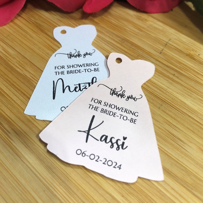 Puede incluir: Dos etiquetas de agradecimiento blancas en forma de vestido con texto negro. La etiqueta superior dice "Gracias por celebrar a la novia" y el nombre "Metz" est&aacute; escrito debajo. La etiqueta inferior dice "Gracias por celebrar a la novia" y el nombre "Kassi" est&aacute; escrito debajo con la fecha "06-02-2024".