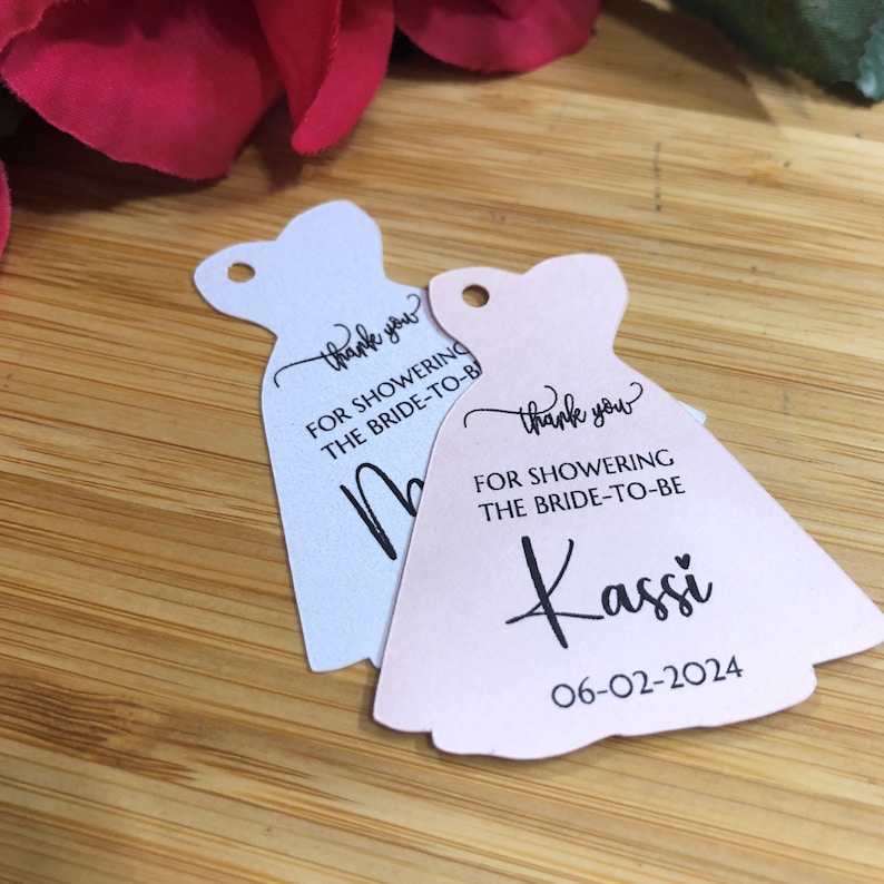 Puede incluir: Dos etiquetas de agradecimiento en forma de vestido, blancas y rosas, para una despedida de soltera. La etiqueta blanca dice "Gracias por celebrar a la futura novia". La etiqueta rosa dice "Gracias por celebrar a la futura novia Kassi 06-02-2024".