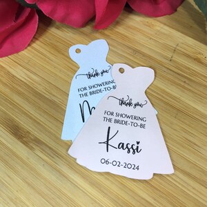 Puede incluir: Dos etiquetas de regalo de despedida de soltera con forma de vestidos. La etiqueta rosa dice "Gracias por celebrar a la novia Kassi 06-02-2024". La etiqueta azul dice "Gracias por celebrar a la novia".