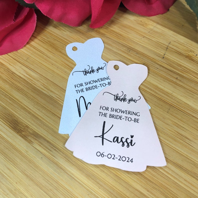 Puede incluir: Dos etiquetas de regalo de despedida de soltera con forma de vestidos. La etiqueta rosa dice "Gracias por celebrar a la novia Kassi 06-02-2024". La etiqueta azul dice "Gracias por celebrar a la novia".