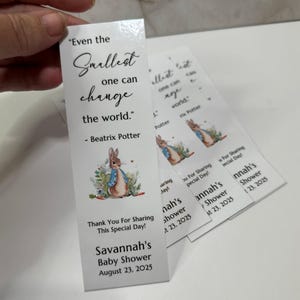 May include: A white rectangular bookmark featuring a watercolor illustration of Peter Rabbit surrounded by greenery. The text reads: "Even the Smallest one can change the world." - Beatrix Potter, "Thank You For Sharing This Special Day!", and "Savannah's Baby Shower August 23, 2025."