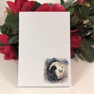 May include: A white card with a black cat sitting on a crescent moon with butterflies flying around it. The image is in a watercolor style.