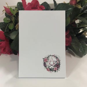 May include: A white notepad with a floral pentacle design. The pentacle is surrounded by pink and purple flowers and leaves.